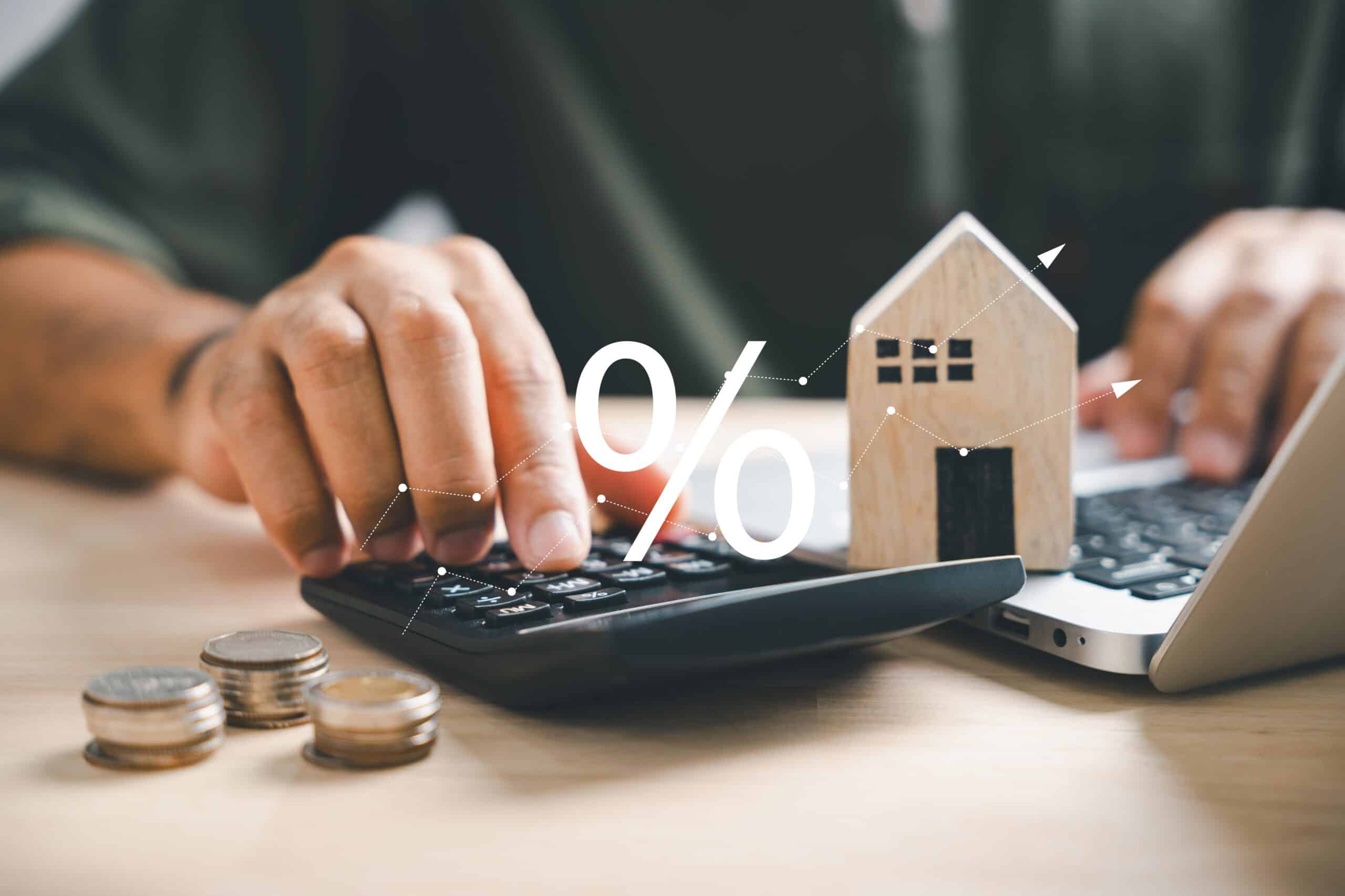 Hands using a calculator and laptop beside a model house and percentage symbol