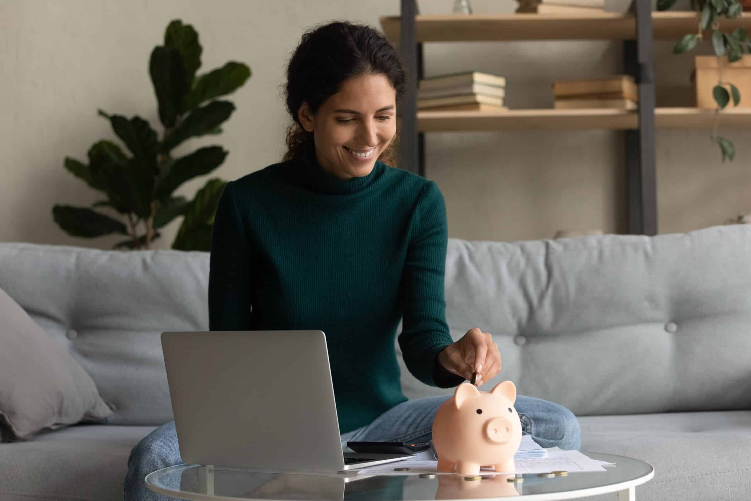 Woman on a sofa with a laptop putting a coin into a piggy bank