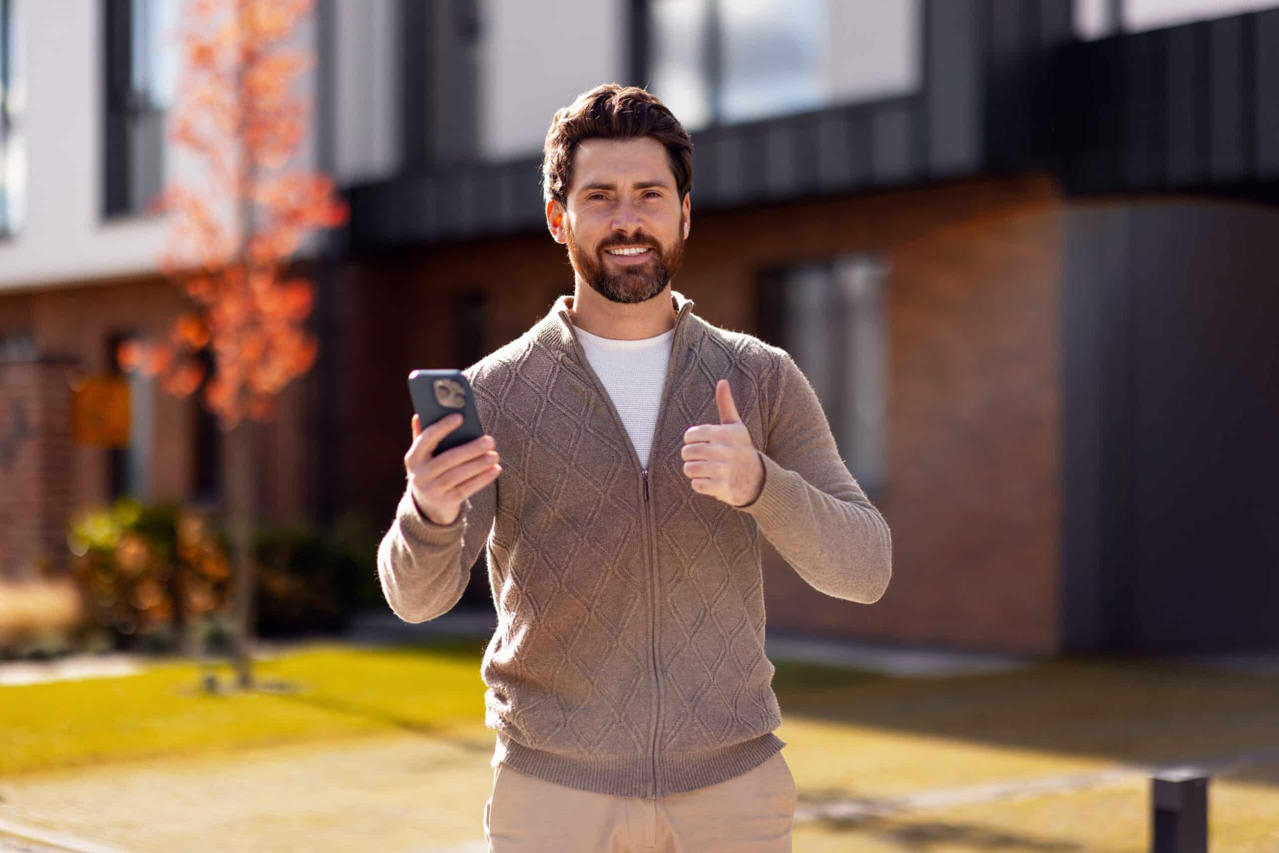 Man outside a modern house holding a phone and giving a thumbs up