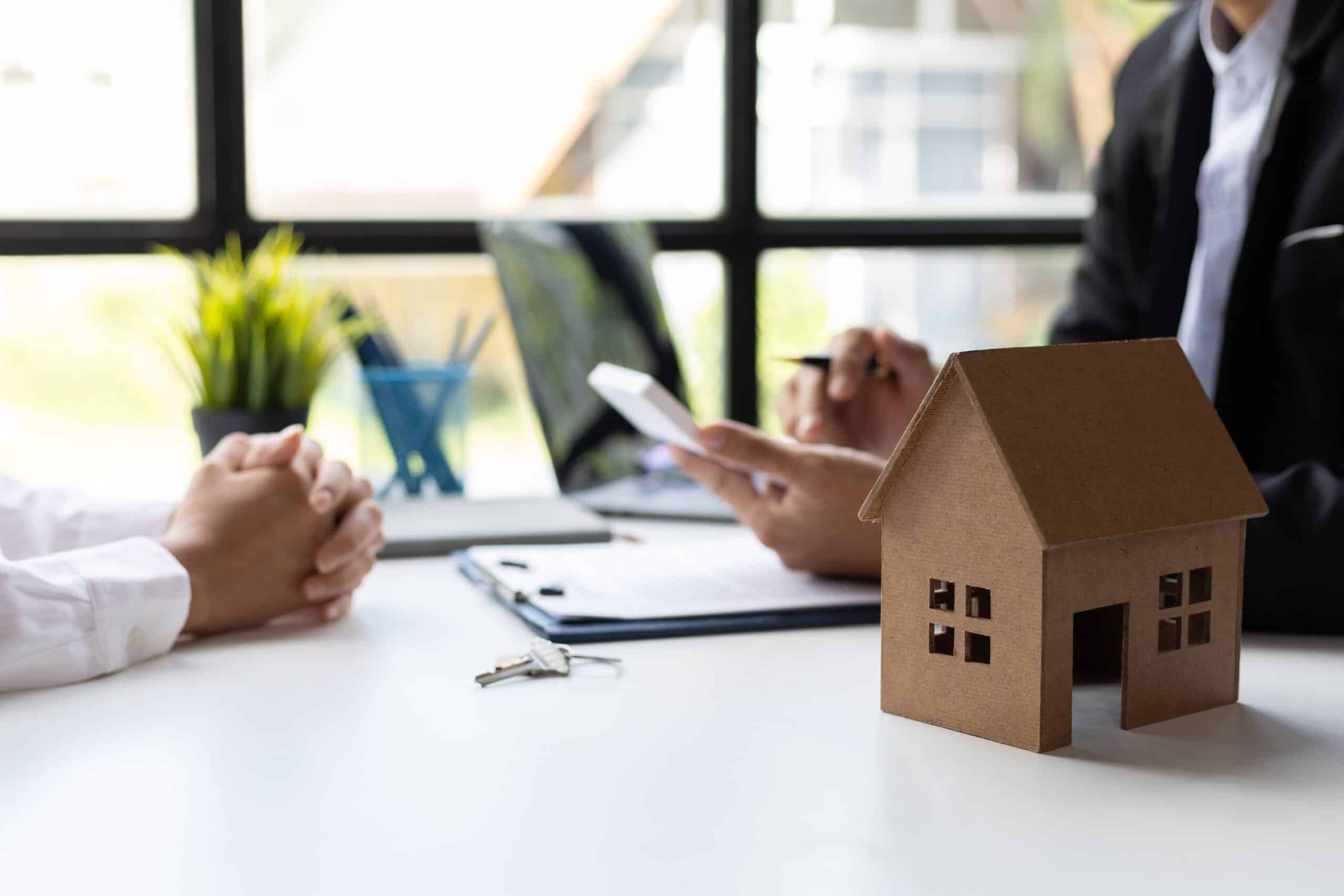 Two people signing paperwork at a desk beside a model house and keys