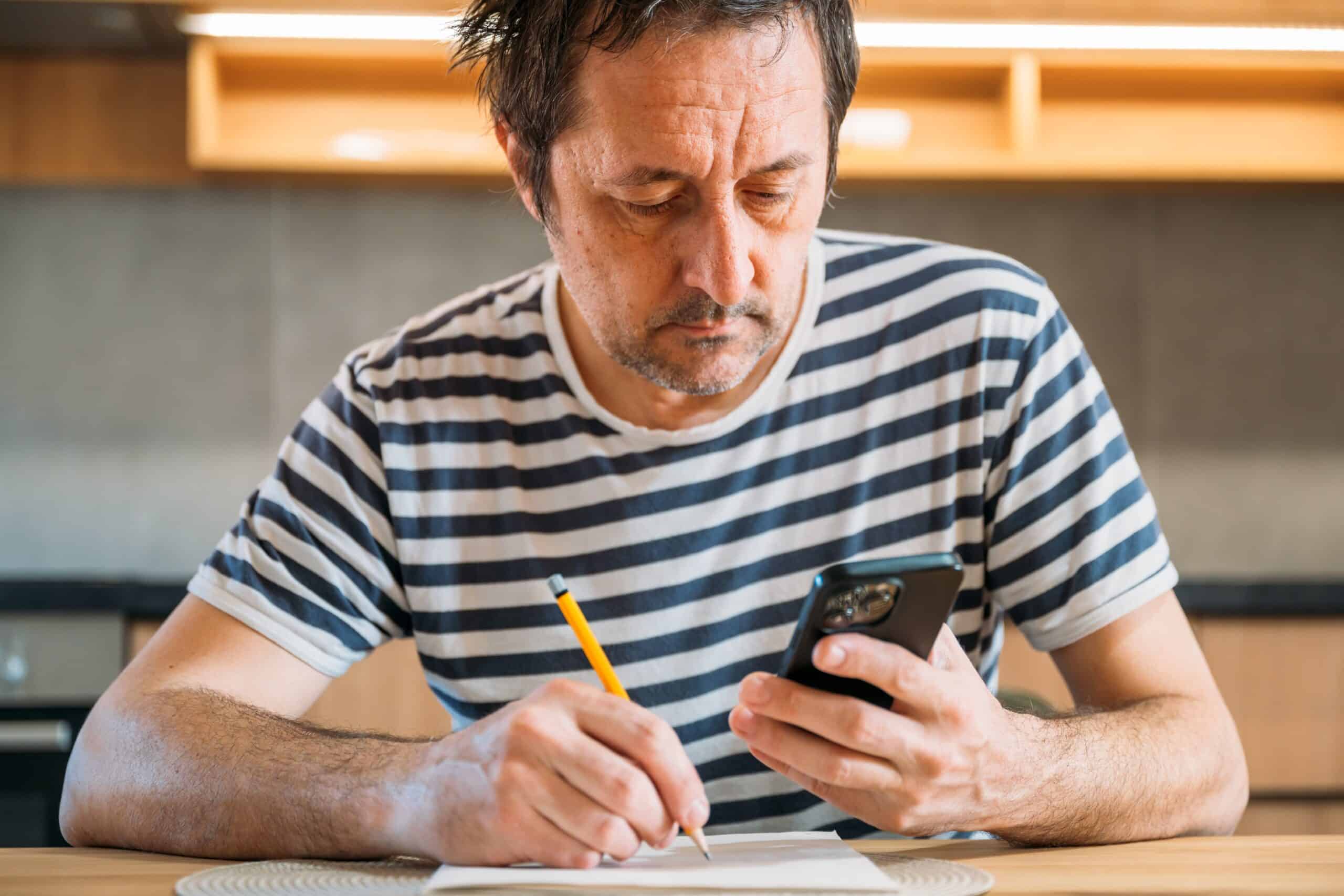 Man writing notes while checking his phone