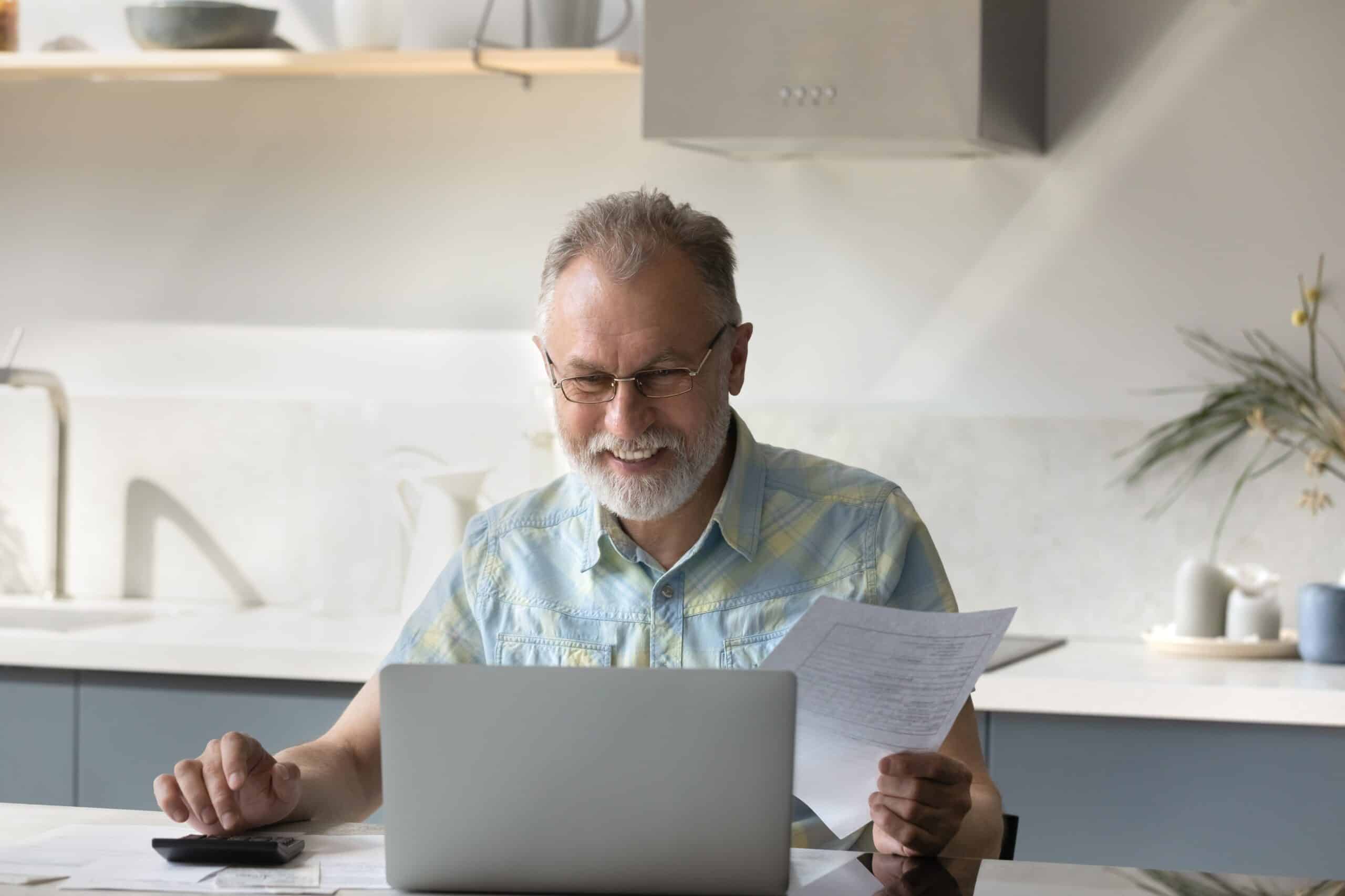 Older man reading a letter at a laptop in a kitchen