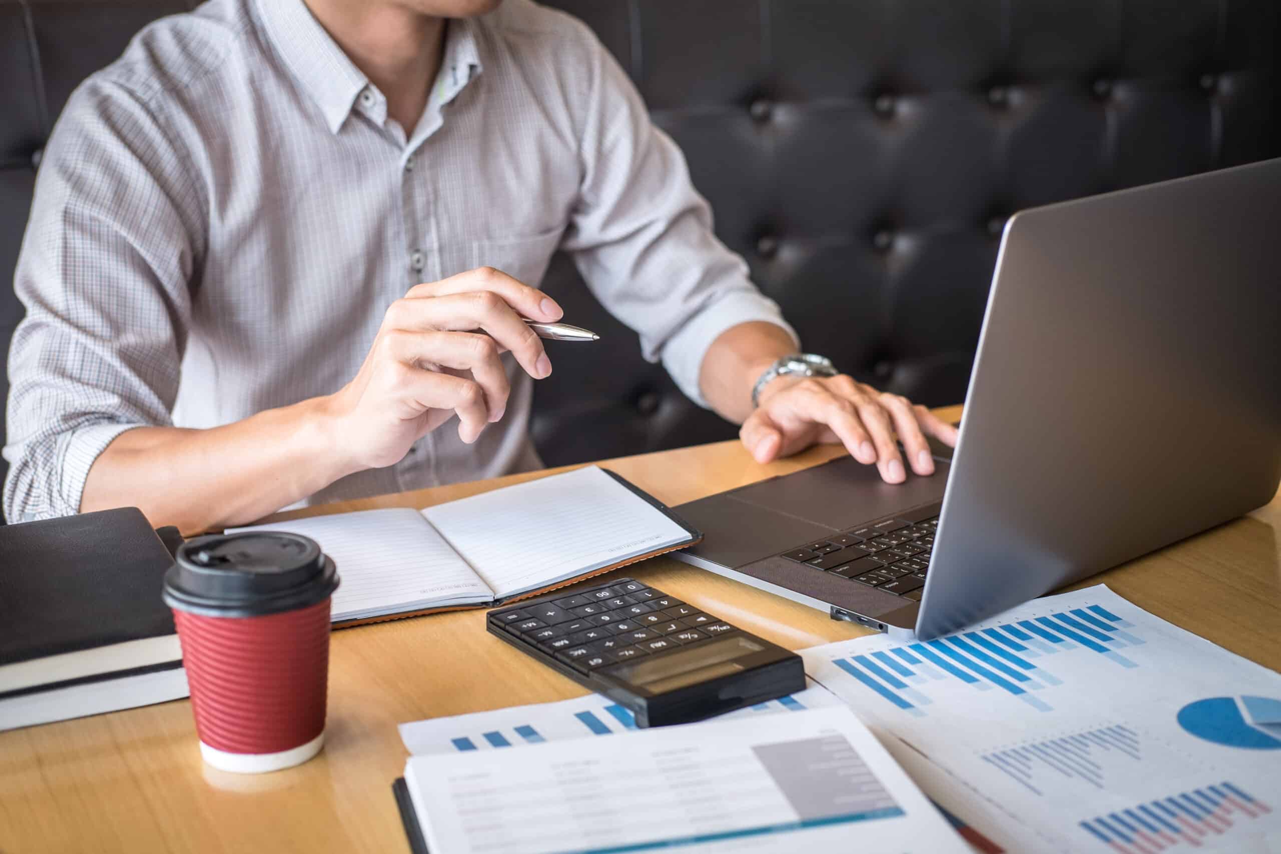 Man working at a laptop with printed charts, a calculator and a coffee cup