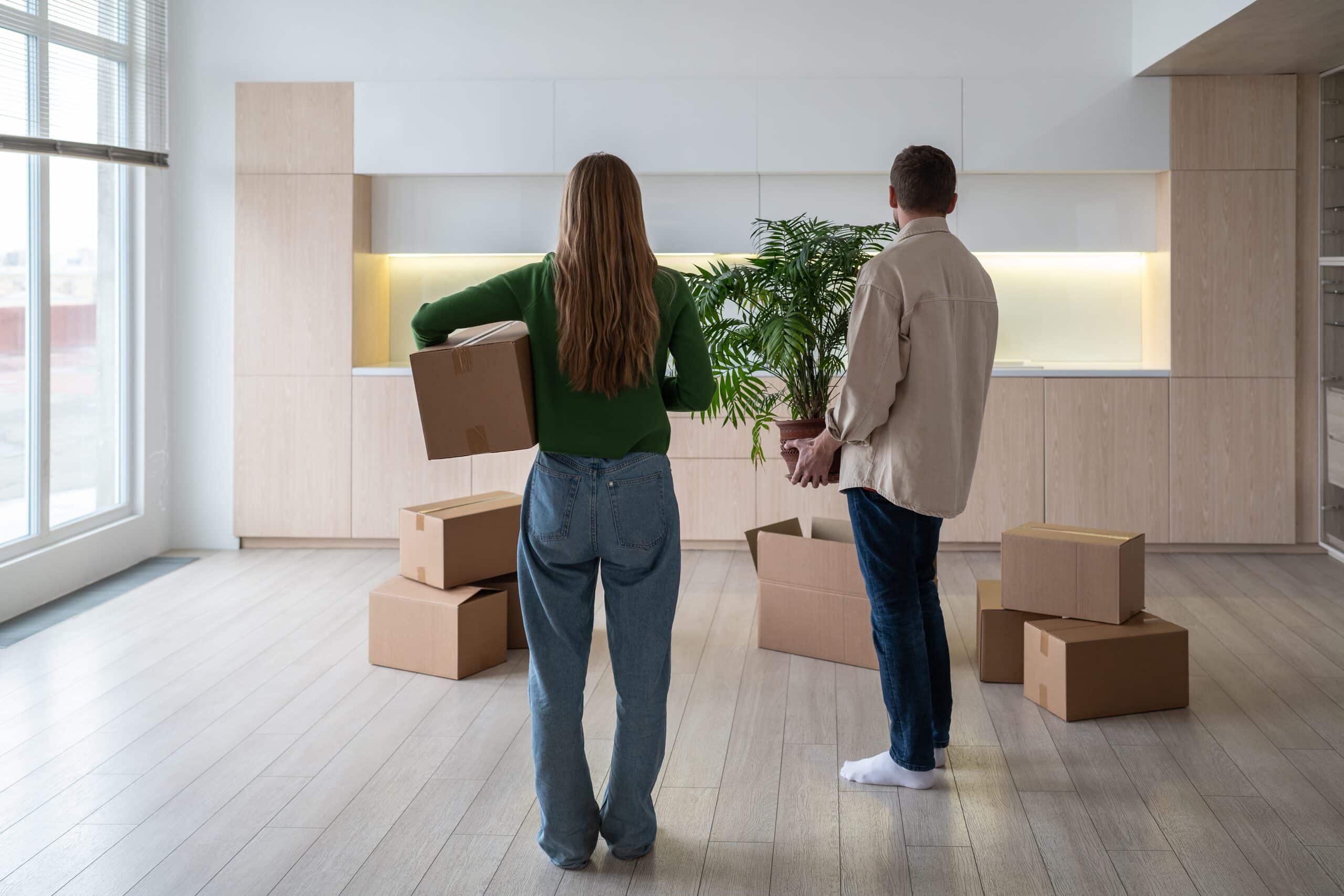 Couple carrying boxes and a plant into an empty flat