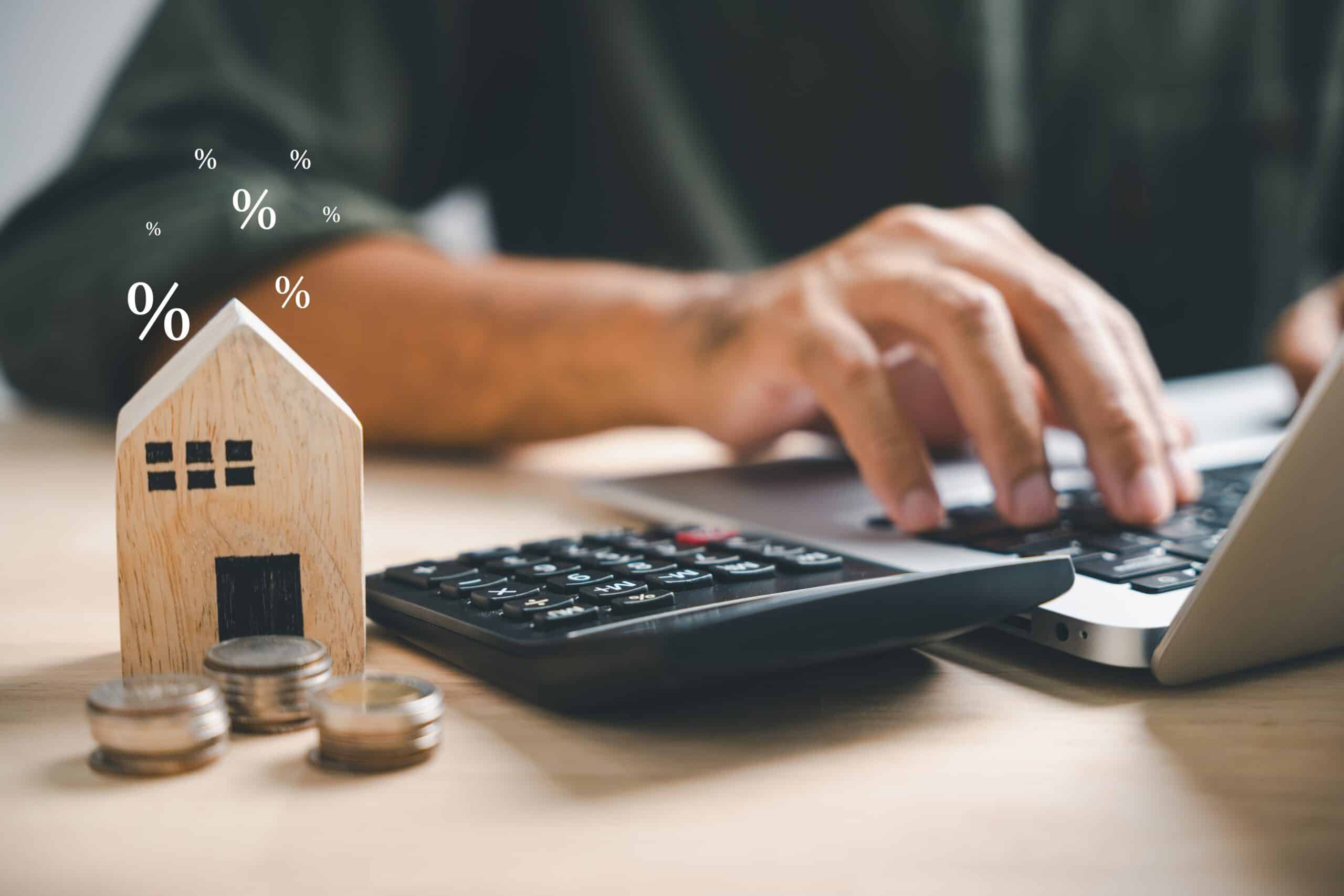 Hands using a calculator beside a wooden model house and percentage symbols